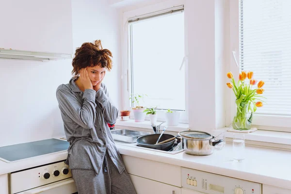 young teenage girl in pajamas, with disheveled hair, with headache, fatigue, depression, stands in kitchen, against blurred background of unwashed dishes, mess. Fatigue and seasonal depression, household chores routine, young lonely woman morning