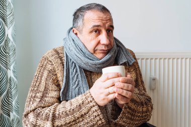 middle-aged man with cold, virus or flu, with runny nose, in warm sweater and woolen scarf, drinks warming drink from cup, an unhappy expression on face. Treatment of colds and flu with vitamin tea
