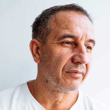 Portrait of middle-aged man with brown eyes, gray hair, mimic wrinkles on face. middle-aged man, with pensive look, sad face, in white T-shirt, close-up, on blurred light background