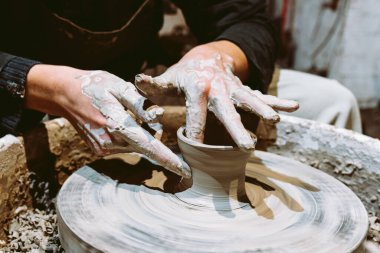 hands young woman close-up are stained in clay, they make ceramic product on potter's wheel. Close-up, soft focus, against blurred background of pottery workshop, Potter's hands at work