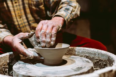 Yakın plan erkek elleri kille lekelenmiş, çömlekçi çarkına çömlek yontuyorlar. Çömlekçilik atölyesinin bulanık arka planına yakın çekim, yumuşak odak. Potter 'ın elleri iş başında.