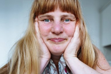 Gülümseyen orta yaşlı, mavi gözlü, sarışın, yüz kırışıklıkları olan, üst dudağında kıllar olan, cildinde kusurlar olan mutlu bir kadın. Yüzü ellerinle yakın çekim yap. Cilt kusurları olan orta yaşlı bir kadının portresi.