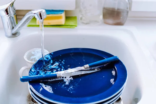 Washing dishes in kitchen sink with sponge. Water flowing from tap onto dirty dishes in kitchen sink. Bright blue plates stacked after breakfast