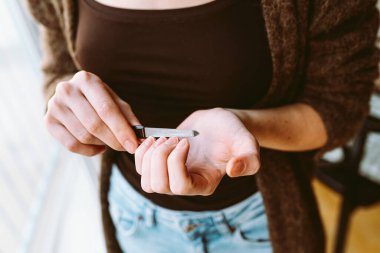 Siyah kozmetik maskeli genç bir kız, boynunda helikopterle, tişörtünde saçı bandajlı, pencerenin yanında duruyor, tırnaklarını törpülüyor. Evde yüz ve ellerle ilgilenmek