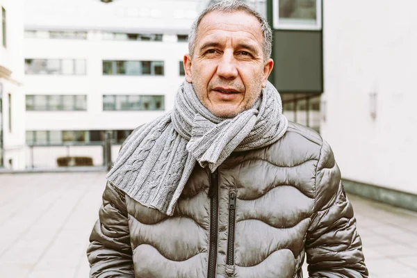 Portrait attractive middle-aged man, with gray short hair, brown-eyed, in warm jacket and scarf, against blurred city background