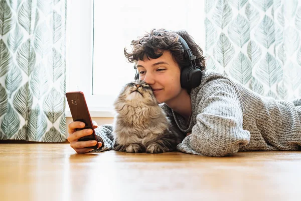 teenage girl in headphones, with mobile phone, lies on floor in living room near large window, with fluffy Maine Coon cat, listens music, spends free time.