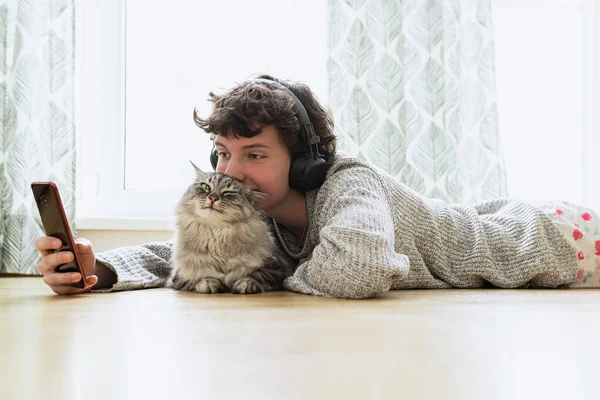 teenage girl in headphones, with mobile phone, lies on floor in living room near large window, with fluffy Maine Coon cat, listens music, spends free time.
