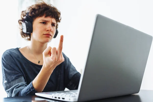 Attractive teenager girl, in headphones, sits in front tablet, shows gesture of middle finger in monitor. concept online communication, self-realization, quarrel, refusal to communicate