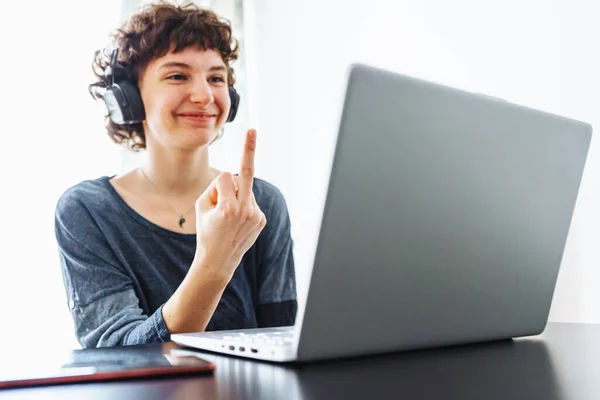 Attractive teenager girl, in headphones, sits in front tablet, shows gesture of middle finger in monitor, with smile. concept online communication, self-realization, quarrel, refusal to communicate