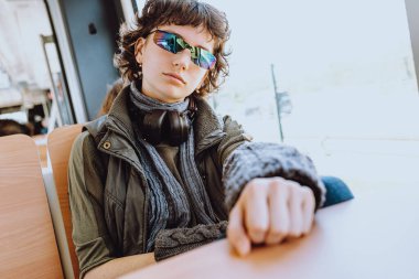 Kulaklıklı hippi genç kız, güneş gözlüğü takıyor, şık kıyafetler giyiyor, müzik dinliyor, tren yolculuğundan hoşlanıyor, seyahat ediyor, pencereden dışarı bakıyor, Z jenerasyonu, yaşam tarzı kavramı.,