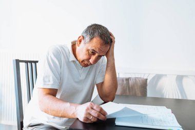 Evrakları tutan, kağıt faturalarını okuyan, emekli maaşlarını, ödemelerini, vergilerini hesaplayan, evde aile emekliliği parası hesaplayan orta yaşlı bir adam. Masada oturan orta yaşlı bir adam fatura okuyor.