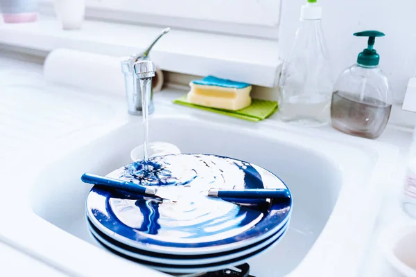 mountain of unwashed dishes in white kitchen sink, water flowing from kitchen faucet. Dirty dishes in sink with water tap and sponge on table