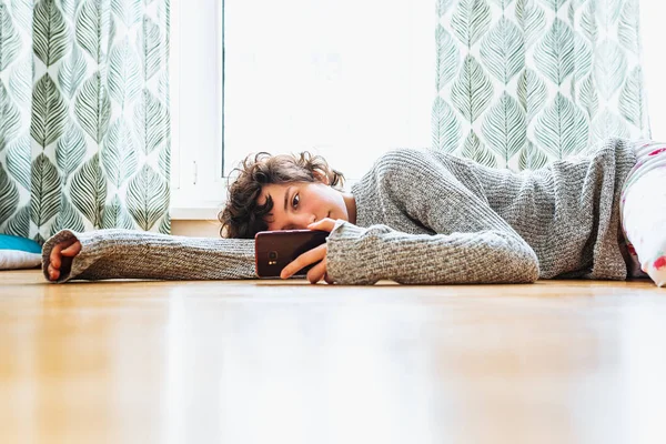 Ev elbiseli genç bir kız, elinde akıllı telefonla kalın gri bir kazak örüyor, parke zeminde yatıyor, yorgun, depresif, kitap okuyor, internette iletişim kuruyor.