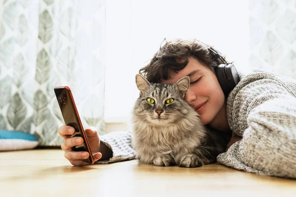 Spend your free time at home with your cat. teenage girl in headphones, with mobile phone, lies on floor in living room near large window, with fluffy Maine Coon cat, listens to music, closing eyes
