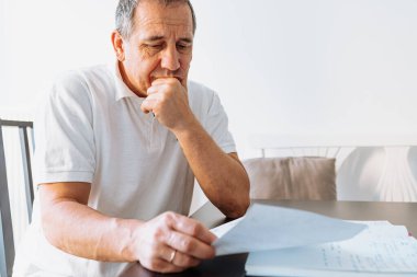 Evrakları tutan, kağıt faturalarını okuyan, emekli maaşlarını, ödemelerini, vergilerini hesaplayan, evde aile emekliliği parası hesaplayan orta yaşlı bir adam. Masada oturan orta yaşlı bir adam fatura okuyor.