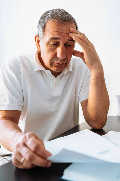 Middle aged man holding documents reading paper bills, calculating pension fees, payments, taxes, planning family retirement money finances at home. middle-aged man at table at home reading bill