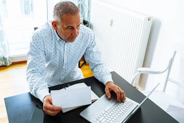 Stock image Middle aged man holding documents reading paper bills, calculator pension fees, payments, taxes, planning family retirement money finances at home. taking notes on laptop