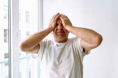 Orta yaşlı bir adam, gri saçlı, stresli, başını tutuyor, baş ağrısı, kayıp, umutsuzluk yaşıyor, beyaz duvara karşı. Erkeklerde orta yaş krizi, suratında asık suratla acı çeken adam portresi.