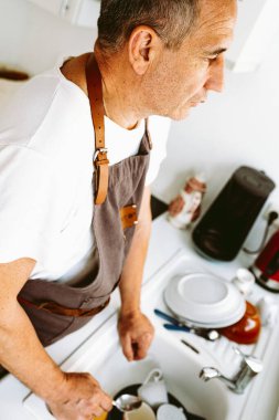 Orta yaşlı bir adam, kaslı, kahverengi mutfak önlüğü giyiyor, mutfakta bulaşık yıkıyor, konsept erkek ev sahibi, ev işleri