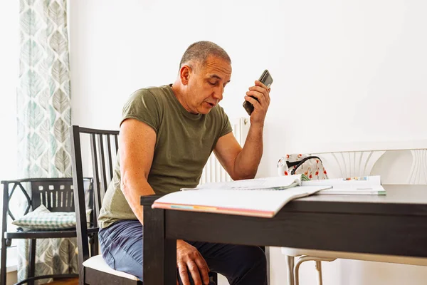 Orta yaşlı bir adam, tişörtlü, kot pantolonlu, oturma odasında masada oturuyor, dizüstü bilgisayar kullanıyor, deftere notlar yazıyor, yabancı diller öğreniyor.