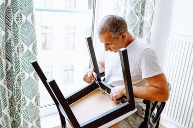 Beyaz tişörtlü çekici, orta yaşlı bir adam tornavidayla sandalyeye vida takıyor, ev mobilyalarını tamir ediyor. Ev işleri, görevler, mobilya tamiri, bozuk ahşap işçiliği.