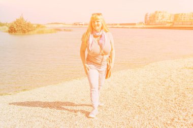 Orta yaşlı bir kadın, uzun sarı saçlı, tombul yapılı, kot pantolonlu, yaz tişörtlü, gök mavisi göle yaz gezisi, güneş gradyanı filtresi