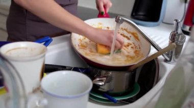 Kahverengi önlüklü genç bir kadın mutfak lavabosunda yanık tavayı, çamaşır bezini ve bulaşık deterjanını yıkıyor. Yakınlarda kirli tabak yığını var..