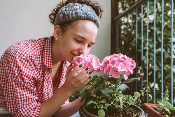 stock image young woman, teenager girl, attractive, in plaid shirt, sits on balcony, terrace, takes care plant in flowerpot, transplants beautiful hydrangea with pink petals