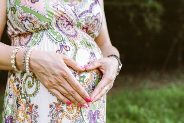 Hamile kadın mideyle kalp şekli yapıyor. Hamilelik, annelik ve yeni aile kavramı. Yeşil Park 'ta güneşli bir yaz gününde kadınların göbek ve kalp sembollerinin yakın çekimi.