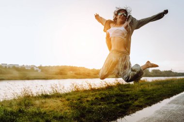 Su birikintilerinde çıplak ayakla dans eden Happy portresi, genç kız, ıslak saçlı, bol yaz kıyafetleri içinde, gün batımında, yağmurdan sonra, nehir kenarında. Doğadan zevk alma kavramı, gün batımı, özgürlük,