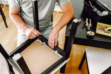 Orta yaşlı bir adam, gri saçlı, kaslı, çekici, sandalyeyi tamir eden, tahta koltukta tornavidayla vidayı sıkan. Ev eşyaları, usta marangoz, ev işleri.