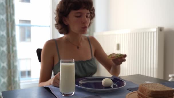 年轻女子 吃土司加鳄梨 煮鸡蛋 喝鲜牛奶 蔬菜牛奶 享受健康的饮食 坐在饭厅的餐桌旁 — 图库视频影像