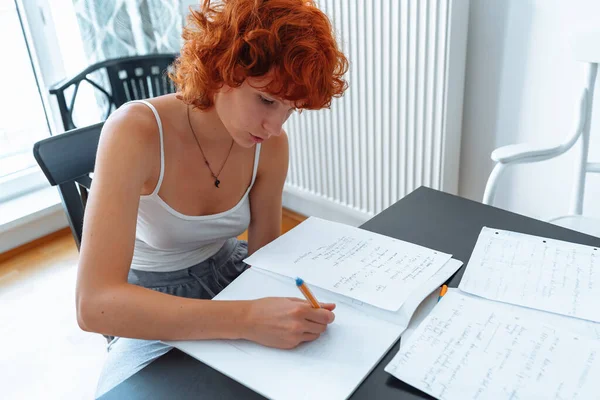 stock image Attractive curly red-haired teenager girl, in T-shirt, at home, sits at table, tucked legs under, studies, writes notes, learns language, self-education