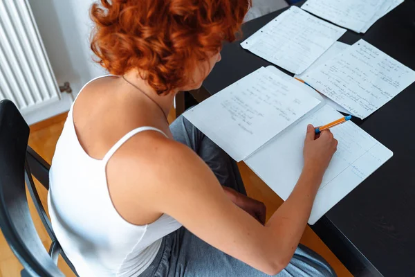 魅力的なカーリー赤毛のティーンエイジャーの少女は Tシャツで 自宅で テーブルに座って 足の下に座り 研究をし ノートを書き 言語を学び 自己教育する — ストック写真