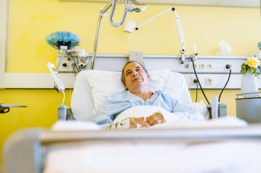 Hasta, orta yaşlı, gri saçlı, mavi gömlekli, modern, uzaktan ayarlanabilir yatakta yatıyor, kolunda kan venöz kateteri var, kolu alçıda, yoğun bakım ünitesinde.