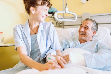 Klinik çalışanları yatalak hastalarla ilgilenir. Orta yaşlı bir adam, kırık kollu, yatağa uzanmış, genç bir kız yanında duruyor, hastaya bakıyor. Hastanedeki hasta bakımı ve bakımı