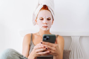 Yüzünde kozmetik çarşaf maskesiyle, kafasında havluyla, açık arka planda, evinde, güzellik salonunda cep telefonu kullanan genç bir kız portresi.
