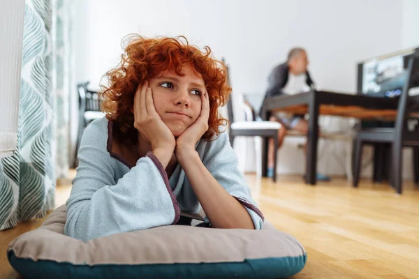 red-haired teenage girl, lying on pillow on floor in living room, with dreamy face, hands under head. Father sits in background, busy, working at computer, watching TV