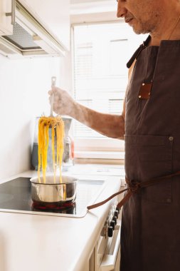 Adam evde, mutfakta makarna pişiriyor. Kahverengi önlüklü bir adam spagettiyi dener, spagettiyi kaynamış suyun üzerinde özel kaşıkta tutar. İtalyan ev yemeği konsepti