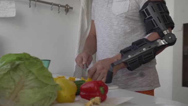 ヒンジ 現代調節可能な肘のOrthosisが付いている堅い肘の整形外科ホールダーが付いている男は サラダ 皮を作り 台所ナイフが付いている野菜を切ります — ストック動画