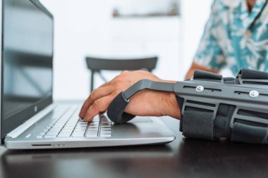 Dirsek yaralanması olan bir adam, dizüstü bilgisayarın klavyesinde özel modern, ayarlanabilir dirsek ortozu yazan bir adam. Yan görüntü, kol bandı takan adam ön camda oturuyor ve evde uzaktan çalışıyor.