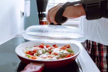 Yakın plan erkek eli, dirsek ekleminden yaralı, modern atel, ortoz, içinde sosis ve sebzelerle omlet pişirilen cam kapaklı kızartma tavasını açar.