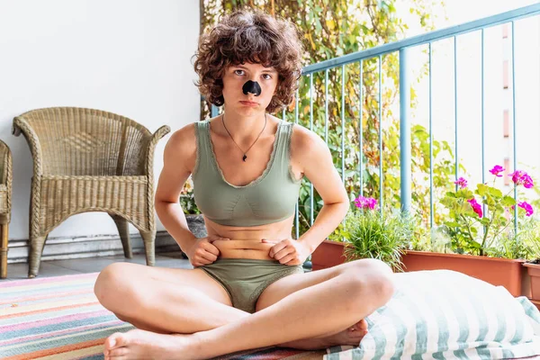 Genç, çekici bir kadın, yeşil bluzlu ve bikinili, burnu açık, kıvırcık saçlı, nilüfer pozisyonunda oturuyor, karnında yağ kıvrımlarını gösteriyor, yoga yapıyor, balkonda ya da terasta.