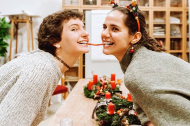 Yeni bir tatil için sosis yiyen komik kız arkadaşların portresi. Farklı uçlardan tütsülenmiş sosis yiyen genç kızların komik portresi, arka plandaki masaya karşı gelişmekte olan çelenklerle.