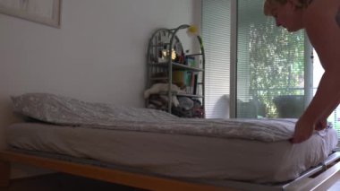 Orta yaşlı, tombul, kilolu, şortlu, tişörtlü, yalın ayaklı, topuz saçlı, yatak odasındaki yatağı toplayan bir kadın.