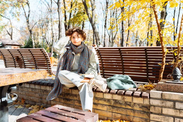 Attractive young woman, wearing scarf, sitting on park bench, holding notepad for notes, smiling, happy