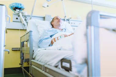 Hasta, orta yaşlı, gri saçlı, mavi gömlekli, modern, uzaktan ayarlanabilir yatakta yatıyor, kolunda kan venöz kateteri var, kolu alçıda, yoğun bakım ünitesinde.