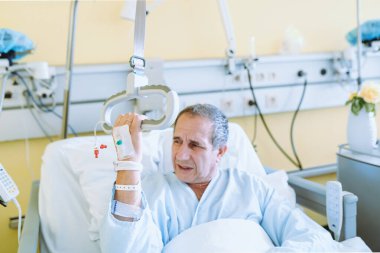 Uyumlu bir hastane yatağında oturan orta yaşlı bir adam yardım çağırmak için korkuluklara tutunuyor. Hastane odası için konfor ekipmanı