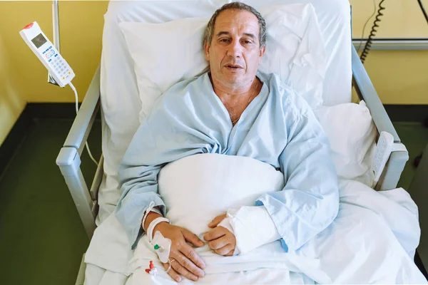 patient, middle-aged man, gray-haired, in hospital blue shirt, lies on modern, remote-adjustable bed, with blood venous catheter on arm, with arm in cast, in intensive care unit hospital