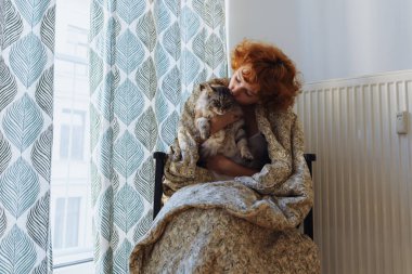 Kucaklayan kedi, battaniye evinde oturan, donmuş. Genç, çıplak ayaklı, kızıl saçlı, kıvırcık saçlı, sabah, battaniyeye sarılmış, ısıtma kaloriferinin yanında oturan evcil kedi, perdelerle kaplı büyük bir pencere..